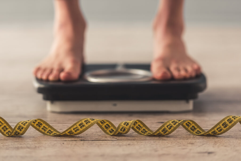 Cropped image of woman feet standing on weigh scales, on gray background. A tape measure in the foreground By VadimGuzhva